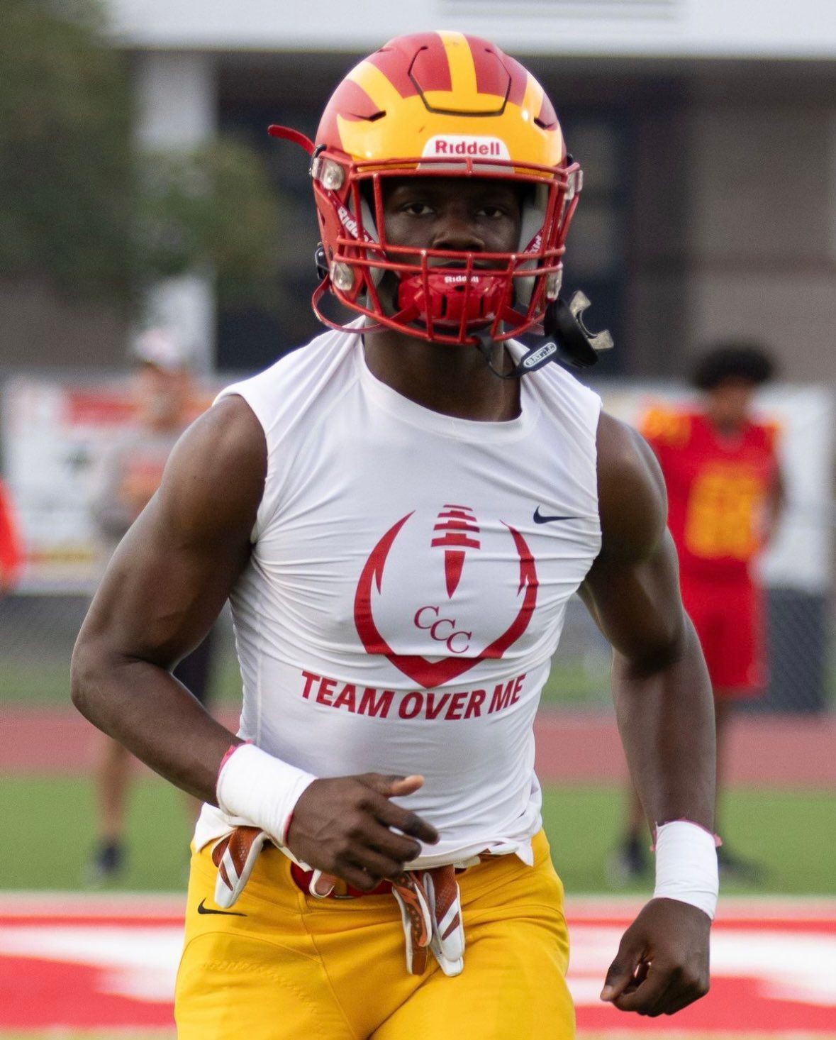 Football player in a red and yellow helmet and white sleeveless shirt on a field during practice.