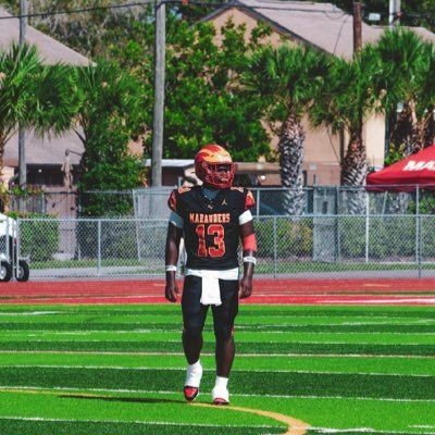 Football player in uniform and helmet standing on a green field with palm trees in the background.