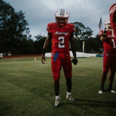 Football player in red uniform on field, wearing helmet, with trees and flag in the background.