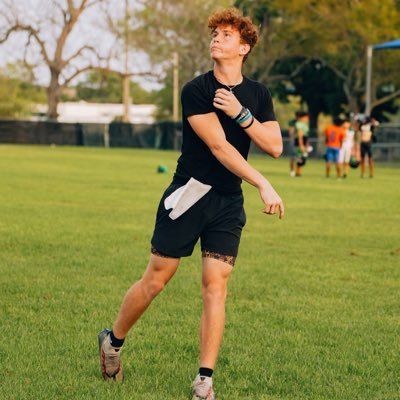 Person in athletic attire playing on a grassy field, with trees and other people in the background.